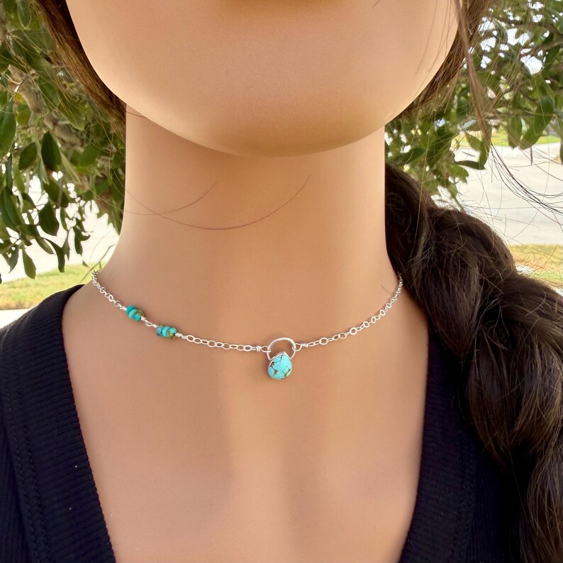 May include: A silver chain necklace featuring a small, teardrop-shaped turquoise pendant suspended from a circular silver link. The chain is accented with three small, round turquoise beads on one side, creating a delicate and balanced design.