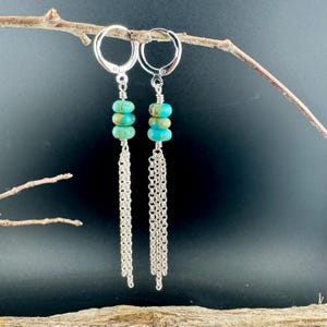 May include: A pair of dangle earrings featuring turquoise-colored stone beads and silver chain tassels. The earrings have a lever-back closure and are suspended from a silver wire. The background is a dark, solid color.