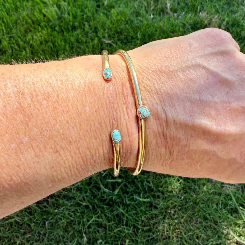 May include: Two gold-colored bracelets with turquoise-colored stones. One is a cuff bracelet, and the other is an open-ended design. The bracelets are worn on a person's wrist, with a green grass background.