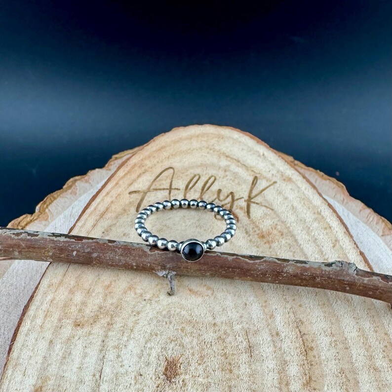K&ouml;nnte beinhalten: Ein silberner Perlenring mit einem schwarzen Stein in der Mitte. Der Ring wird auf einer Holzoberfl&auml;che mit dem Text "Ally k" im Hintergrund pr&auml;sentiert. Ein schlichtes und elegantes Schmuckst&uuml;ck.