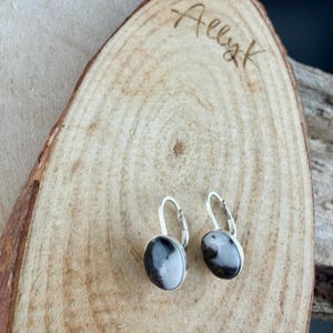 May include: A pair of oval-shaped earrings with a silver-colored metal setting. The earrings feature a black and white stone. The earrings are displayed on a wooden surface with the handwritten text "Allyk".