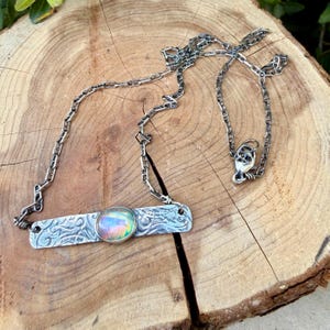 May include: A silver-toned necklace featuring a rectangular metal bar pendant with an embossed floral pattern and a central oval iridescent cabochon stone. The pendant is attached to a delicate silver-toned chain with a lobster claw clasp.