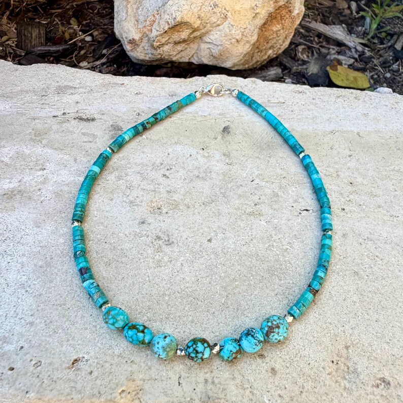 May include: Turquoise beaded necklace with a silver clasp. The necklace features a mix of small, flat beads and larger, round beads in shades of blue and green. The necklace is approximately 16 inches long.