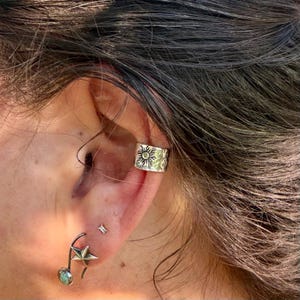 May include: A silver ear cuff featuring a textured sun motif worn on the upper cartilage, paired with a silver stud earring shaped like a star and a small dangling circular stone charm attached to a curved metal wire on the earlobe.