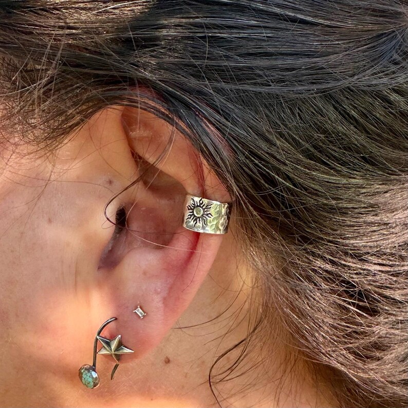 May include: A silver ear cuff featuring a textured sun motif worn on the upper cartilage, paired with a silver stud earring shaped like a star and a small dangling circular stone charm attached to a curved metal wire on the earlobe.