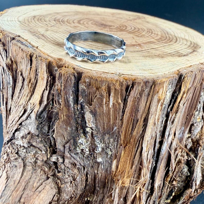 May include: A silver-toned metal ring featuring a continuous, repeating wave-like pattern etched along the outer band. The ring has a polished finish with darkened recessed areas that highlight the textured, undulating design around the circumference.