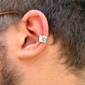 May include: A silver-colored ear cuff featuring a hammered, textured surface and a stamped sun design. The metal band is open-ended and wraps around the cartilage of the ear, displaying a polished finish with a detailed celestial motif.