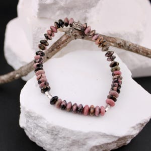 May include: A beaded bracelet featuring pink and black gemstone beads. The bracelet is strung on a flexible cord and has a silver-colored clasp. The beads are oval-shaped and vary in size. The bracelet is displayed on a white surface.