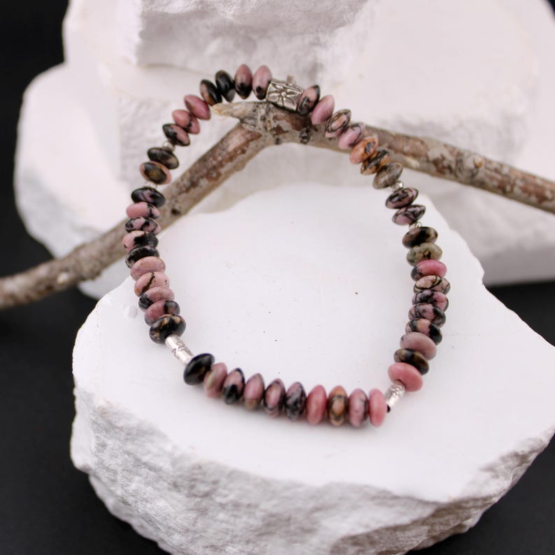 May include: A beaded bracelet featuring pink and black gemstone beads. The bracelet is strung on a flexible cord and has a silver-colored clasp. The beads are oval-shaped and vary in size. The bracelet is displayed on a white surface.