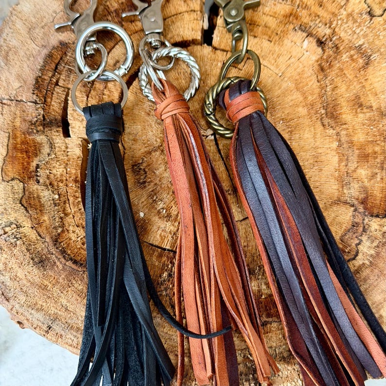 May include: Three leather tassel keychains in black, tan, and brown. Each keychain has a metal clasp and a ring to attach keys. The tassels are made of strips of leather and are tied at the top. The keychains are displayed on a wooden surface.