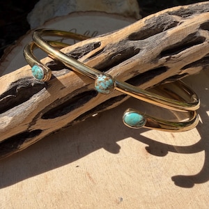 May include: A gold-colored cuff bracelet with three turquoise stones. The bracelet has a curved design and is displayed on a piece of driftwood. The turquoise stones are round and set in gold-colored metal.