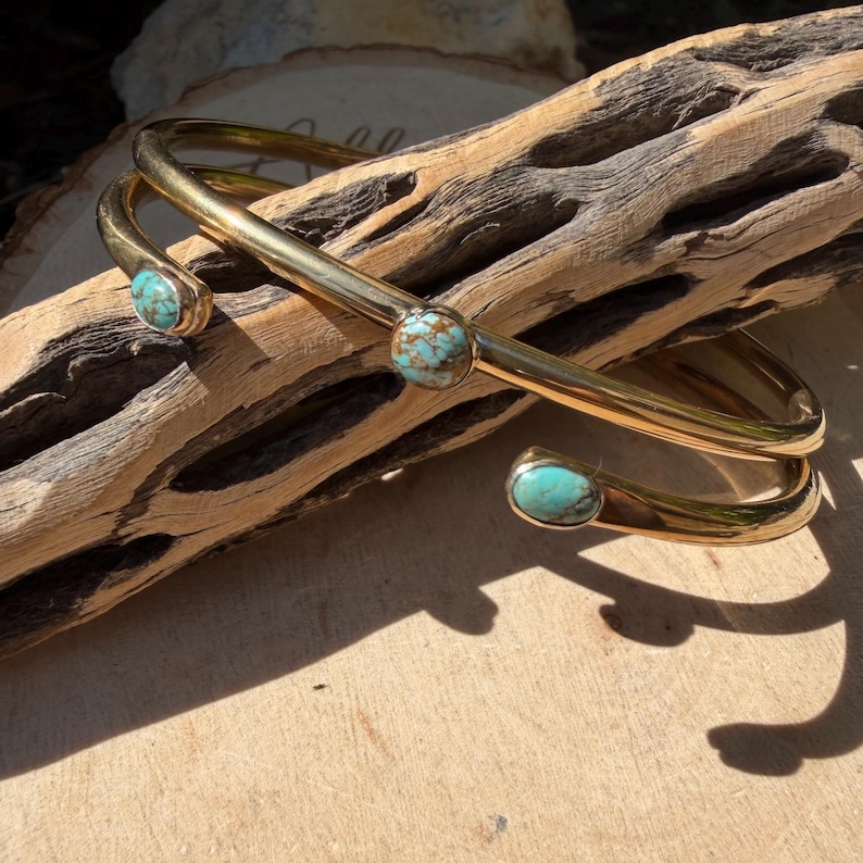 May include: A gold-colored cuff bracelet with three turquoise stones. The bracelet has a curved design and is displayed on a piece of driftwood. The turquoise stones are round and set in gold-colored metal.
