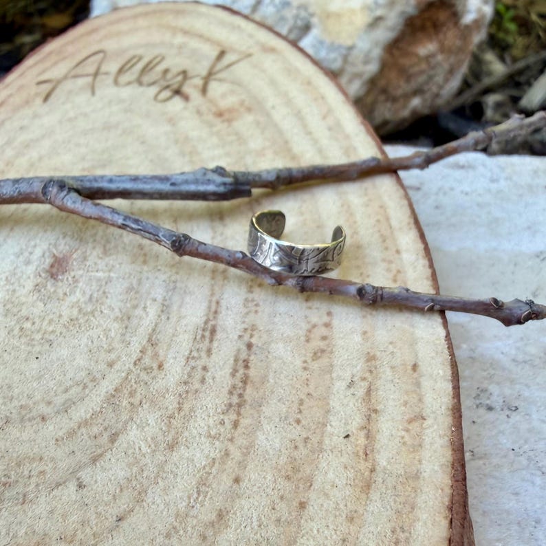 May include: A silver-colored adjustable ring with a textured surface, resting on a small twig. The ring is open at the bottom and sits on a light brown wooden surface. The word "Ally K" is written on the wood.