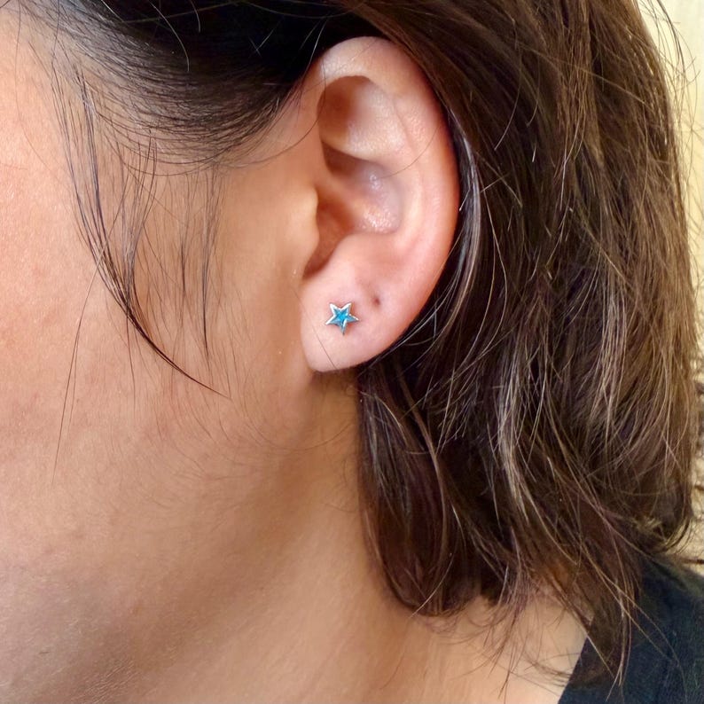 May include: A close-up of a person's ear with a small, silver star-shaped earring with a turquoise center. The earring is a stud style. The person has dark hair and is wearing a black shirt.