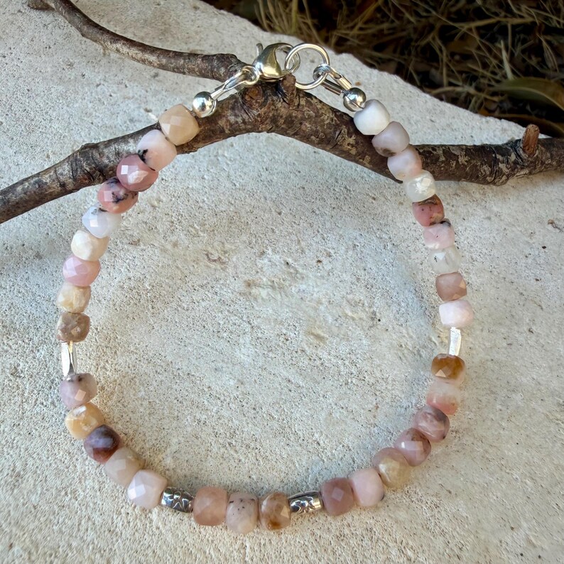 May include: A beaded bracelet featuring faceted, multi-colored gemstone beads in shades of pink, peach, and cream. The bracelet includes small silver-toned metal spacers and a silver-toned lobster claw clasp, creating a delicate and circular jewelry piece.