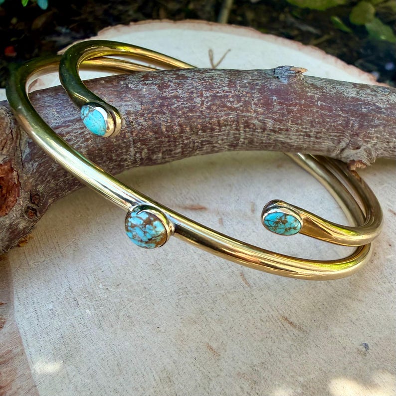 May include: Two gold-colored open cuff bracelets with turquoise stones. The bracelets are resting on a piece of wood. The turquoise stones have a mottled appearance with shades of blue and brown. The bracelets have a smooth, polished finish.