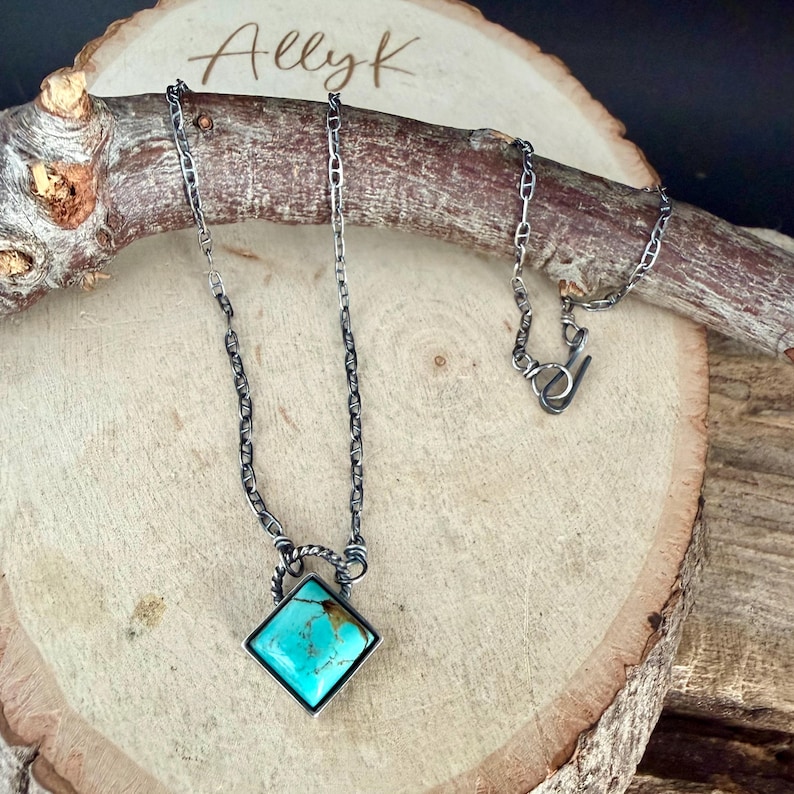 May include: A silver necklace with a square turquoise pendant. The pendant is set in a silver frame and hangs from a chain. The necklace is displayed on a wooden surface with the name "Allyk" written on it.
