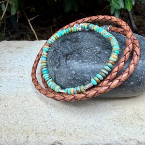 May include: A multi-strand bracelet featuring a braided brown leather cord and a strand of small, round, turquoise-colored stone beads. The beads are interspersed with small, polished silver-toned metal spacers, and the bracelet is secured with a silver-toned metal clasp.