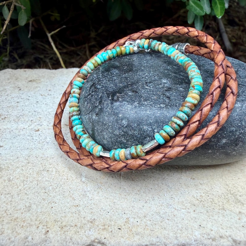 May include: A multi-strand bracelet featuring a braided brown leather cord and a strand of small, round, turquoise-colored stone beads. The beads are interspersed with small, polished silver-toned metal spacers, and the bracelet is secured with a silver-toned metal clasp.