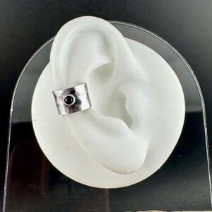 May include: A silver ear cuff with a hammered texture and a small, round black stone. The cuff is displayed on a white ear model against a dark background. This piece of jewelry is designed to be worn on the ear.
