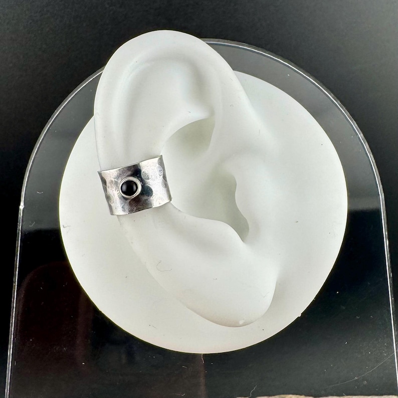 May include: A silver ear cuff with a hammered texture and a small, round black stone. The cuff is displayed on a white ear model against a dark background. This piece of jewelry is designed to be worn on the ear.