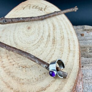 May include: A silver ring with a hammered texture and a round, purple gemstone. The ring is displayed on a wooden stick, set against a wooden background. The image is signed "Ally K" in the upper left corner.