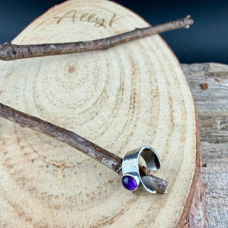 May include: A silver ring with a hammered texture and a round, purple gemstone. The ring is displayed on a wooden stick, set against a wooden background. The image is signed "Ally K" in the upper left corner.