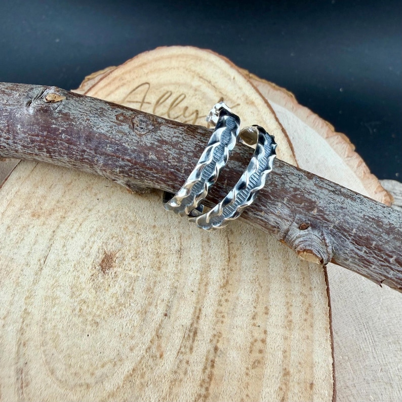May include: Two silver-colored hoop earrings with a textured, wave-like design. The earrings are displayed on a wooden branch, set against a wooden background. The earrings have a dark, oxidized finish in the grooves.