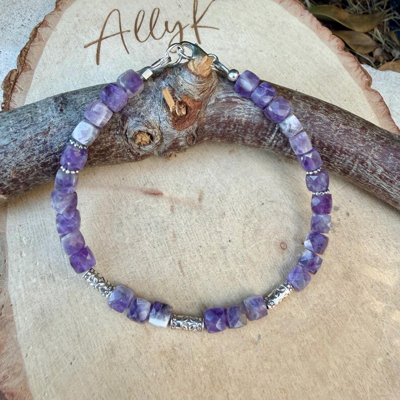 Puede incluir: Una pulsera de cuentas compuesta por cubos de amatista morada facetada intercalados con peque&ntilde;as cuentas espaciadoras de metal plateado. La pulsera se asegura con un cierre de mosquet&oacute;n plateado y se muestra sobre una superficie de madera clara.