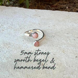 May include: A silver open-style ring featuring two round, light pink stones, each measuring 5mm. The ring has a textured, hammered band and smooth bezels holding the stones in place. The text 5mm stones, smooth bezel & hammered band is visible.