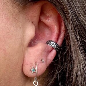 May include: Close-up of an ear adorned with jewelry. A silver ear cuff with a floral design is on the upper ear. A star-shaped earring with a turquoise center hangs from the earlobe. The jewelry is set against the backdrop of skin and hair.