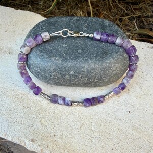 Puede incluir: Una pulsera con cuentas de amatista facetadas en forma de cubo en tonos morados y blancos, intercaladas con peque&ntilde;as cuentas espaciadoras de metal plateado. La pieza se asegura con un cierre de mosquet&oacute;n y una anilla de color plateado.