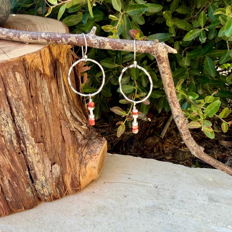 May include: A pair of silver-toned hoop earrings featuring a textured, irregular circular frame. Each earring is accented with a vertical drop consisting of two small, cylindrical, coral-colored beads separated by a tiny silver-toned spacer bead.