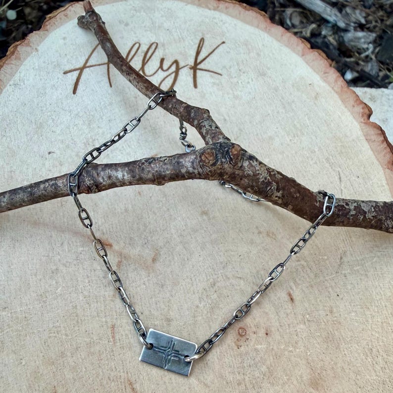 May include: A silver-toned necklace with a rectangular pendant featuring a cross design. The necklace is composed of a chain with elongated links. The jewelry is displayed on a wooden surface with the handwritten text "Allyk".