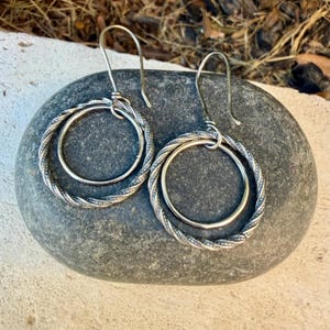 May include: A pair of silver-toned earrings featuring a double-hoop design. Each earring consists of a smaller smooth inner circle suspended within a larger, twisted rope-textured outer circle, attached to a curved ear wire hook.