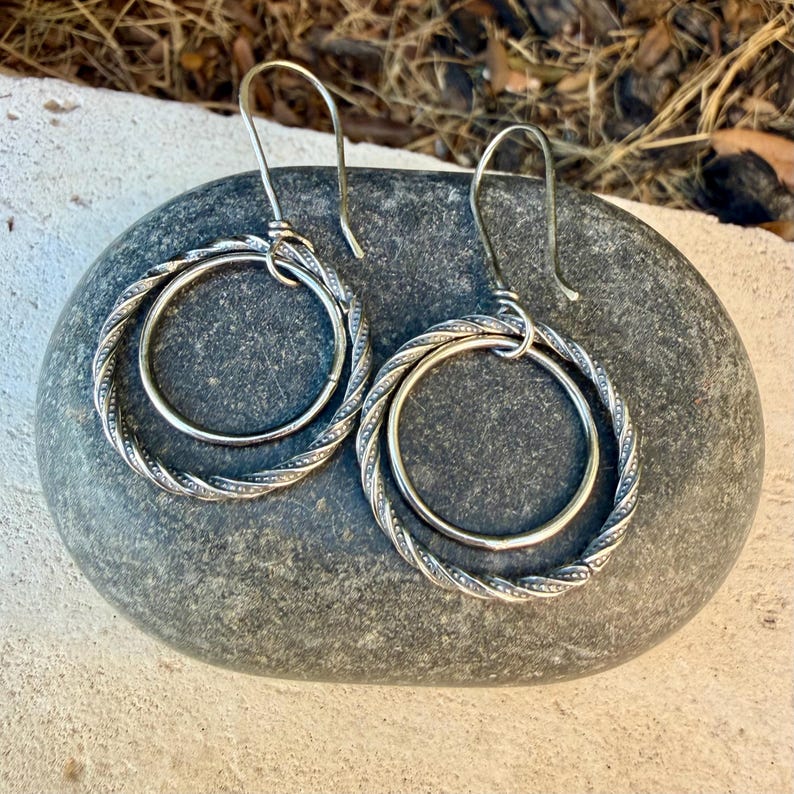 May include: A pair of silver-toned earrings featuring a double-hoop design. Each earring consists of a smaller smooth inner circle suspended within a larger, twisted rope-textured outer circle, attached to a curved ear wire hook.