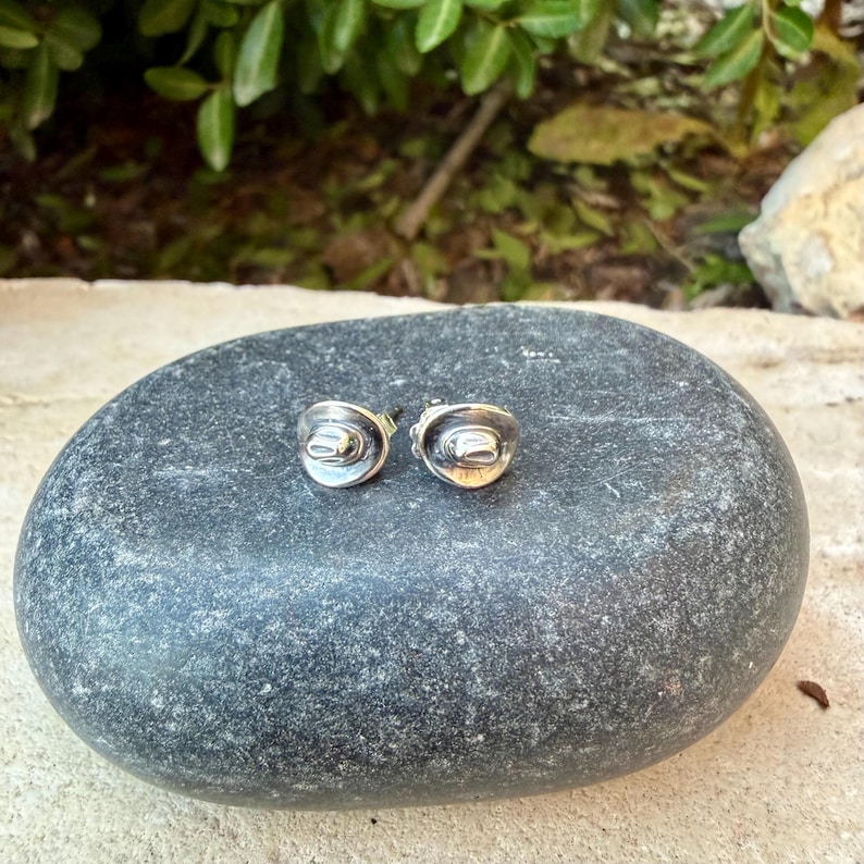 May include: A pair of silver-coloured stud earrings in the shape of miniature cowboy hats. The earrings are displayed on a smooth, dark grey stone, with a blurred green background.