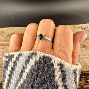 May include: A silver ring with a black diamond-shaped stone. The band has a braided design. The ring is on a finger, with a gray and white patterned sweater sleeve in the foreground. The background is a wooden surface.