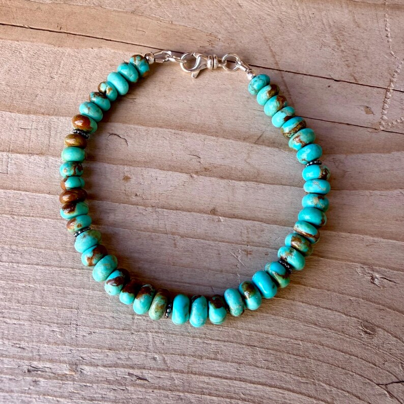May include: A bracelet composed of round, turquoise-colored beads with brown matrix inclusions. The beads are strung together and secured with a silver-toned metal lobster claw clasp. The bracelet forms a circular shape against a wooden surface.