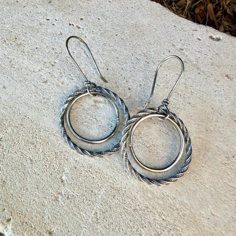May include: A pair of silver-toned earrings featuring a double-hoop design. Each earring consists of a smooth inner circle suspended within a larger, twisted rope-textured outer circle, attached to a curved ear wire hook for secure placement.