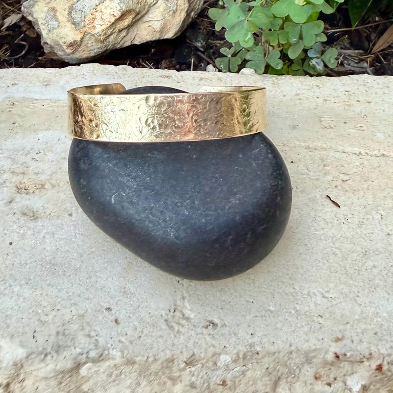 Puede incluir: Un brazalete tipo pu&ntilde;o de metal dorado que presenta una banda ancha con un acabado de superficie martillado y texturizado. El brazalete tiene un dise&ntilde;o abierto, lo que permite colocarlo en la mu&ntilde;eca, y muestra un brillo met&aacute;lico reflectante.