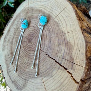 May include: A pair of dangle earrings featuring teardrop-shaped turquoise stones set in silver. Each earring has a single stone at the top with two long, thin, textured silver metal wires extending downward, ending in small rounded silver tips.