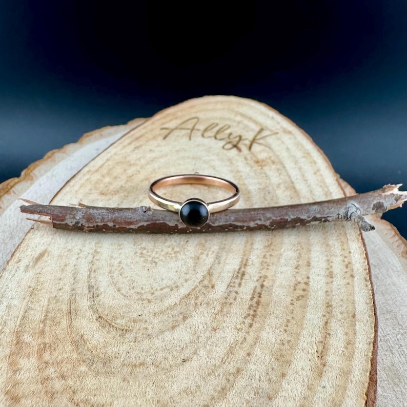 May include: A rose gold ring featuring a round, black gemstone. The ring is presented on a small twig, placed on a wooden surface. The name "Ally K" is inscribed on the wood.