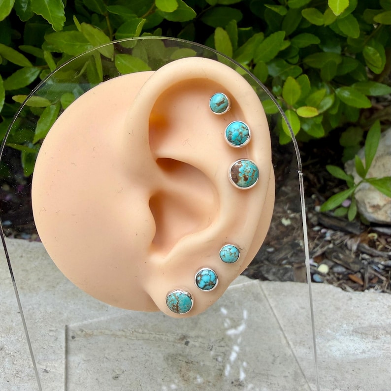 May include: A display of turquoise stud earrings in various sizes, set in silver. The earrings are arranged on a realistic ear model, showcasing the different placements for ear piercings. The turquoise stones have a natural, mottled appearance.