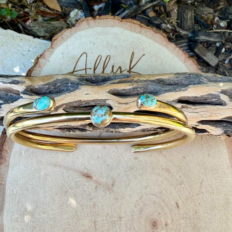 May include: A gold-colored cuff bracelet with three turquoise-colored stones. The bracelet is displayed on a piece of wood with the word "Alluk" written on a wooden slice in the background. The bracelet has a simple, elegant design.
