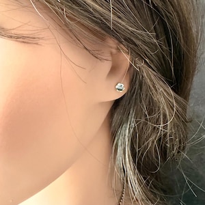 May include: A small, silver-toned stud earring shaped like a four-leaf clover. The earring features a polished metallic finish and is secured directly against the earlobe, displaying a simple and compact design with four rounded petals.