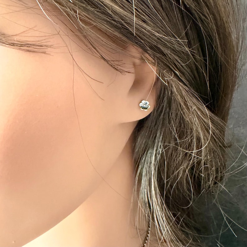 May include: A small, silver-toned stud earring shaped like a four-leaf clover. The earring features a polished metallic finish and is secured directly against the earlobe, displaying a simple and compact design with four rounded petals.