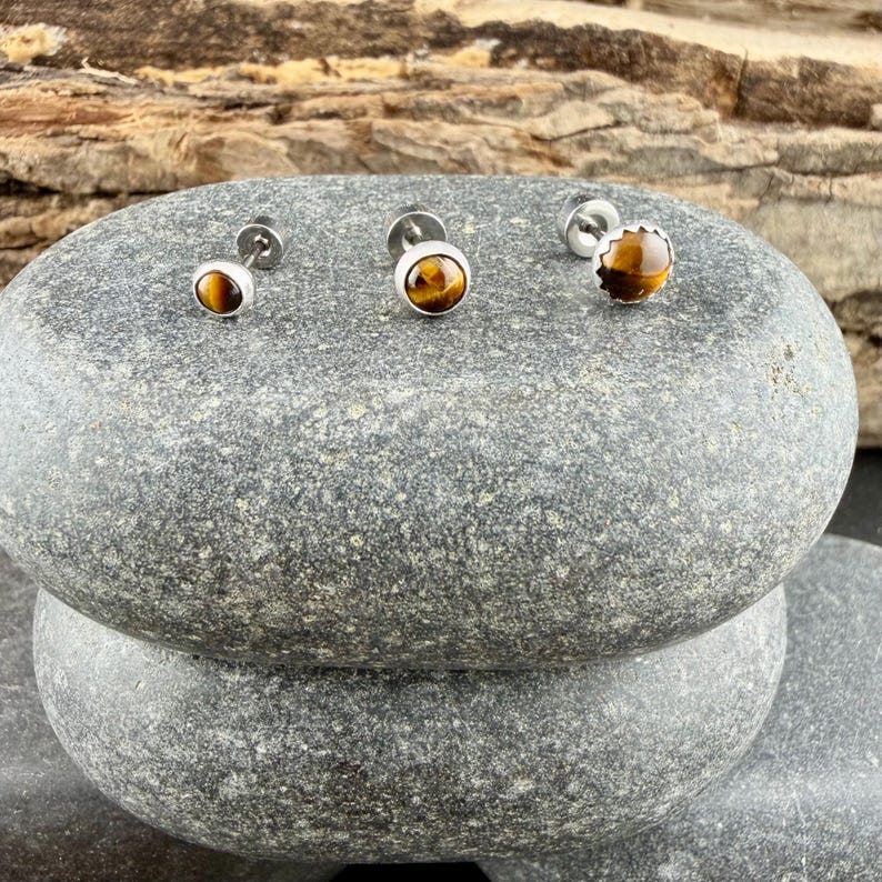 Puede incluir: Tres pendientes de bot&oacute;n con piedras preciosas de ojo de tigre redondas y pulidas, engastadas en metal plateado. Cada pendiente presenta una piedra lisa en corte cabuj&oacute;n con bandas de color marr&oacute;n dorado y brillo.