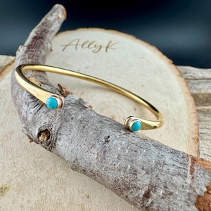 May include: A gold-coloured metal cuff bracelet with turquoise-coloured stones at each open end. The bracelet is resting on a piece of wood. The name "Allyk" is written on the wood.