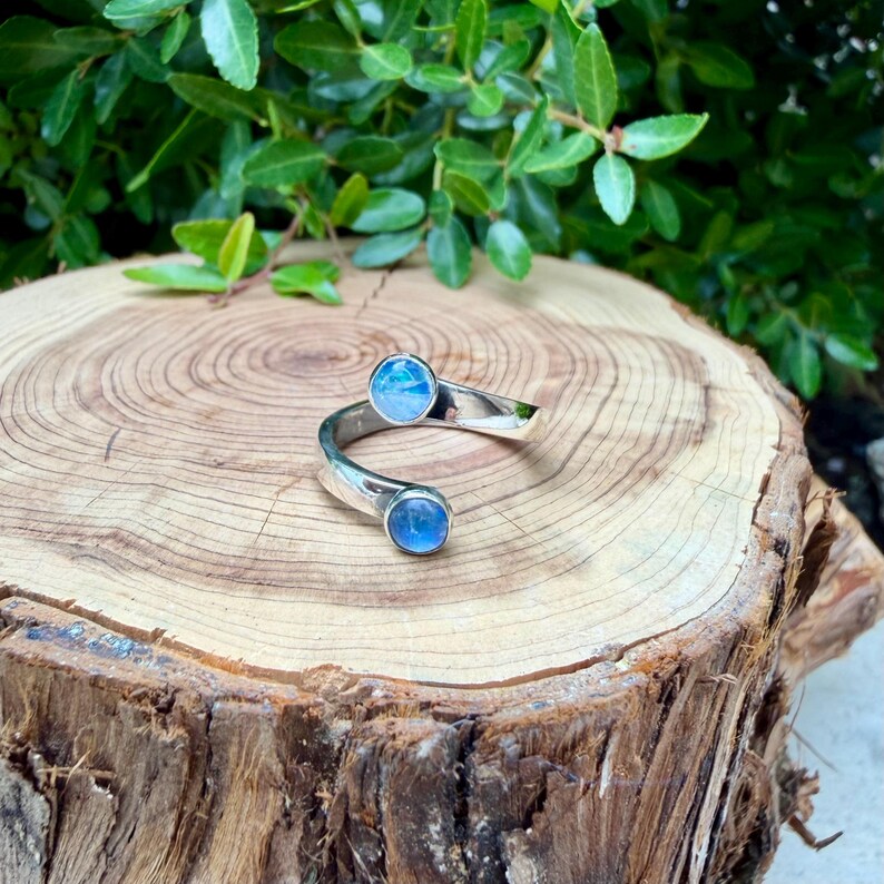 May include: A silver ring featuring two round, blue gemstones. The ring has an open, swirling design, with the gemstones set at either end of the band. The ring is displayed on a wooden surface with green foliage in the background.